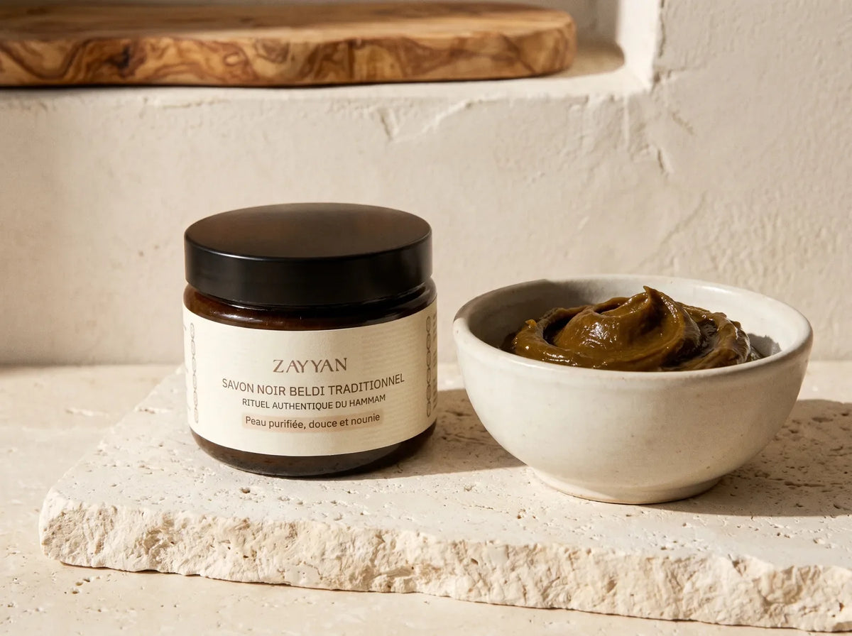 Pot de savon noir Zayyan fermé sur une pierre calcaire crème, à côté d'un bol de pâte noire