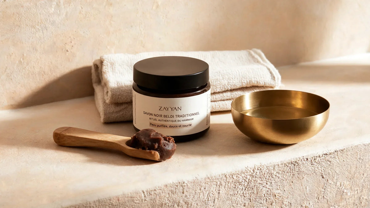 Pot de savon noir beldi Zayyan sur un rebord en tadelakt, une spatule en bois d'olivier chargée de pâte brune, bol de laiton et linge plié